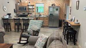 A cozy living room with a brown sofa, a wooden rocking chair, and a coffee table. A kitchen with wooden cabinets, a microwave, and a refrigerator is visible in the background.