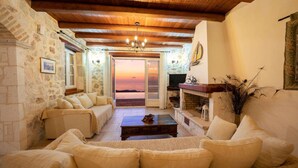 A cozy living room with stone walls, wooden beams, a sofa set, a fireplace, and a view of the sunset. - Impressive Stone Villa, A/C, Private Pool, Sweeping Sea Views to Assos Bay & Magnificent Sunsets! (Defaranata)