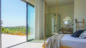 Ein Schlafzimmer mit einer großen Schiebeglas-Tür, die auf einen Balkon führt, einem Bett mit Hängesessel, einem hölzernen Nachttisch, einem runden Spiegel und Blick auf eine hügelige Landschaft.