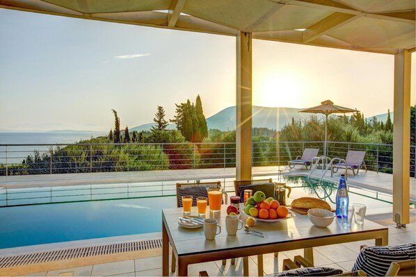 A poolside dining area with a table set for breakfast, overlooking a scenic view of the sea and mountains.