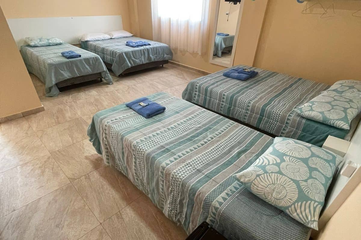 A hotel room with four beds, a small black refrigerator, a mirror, and a window with curtains.