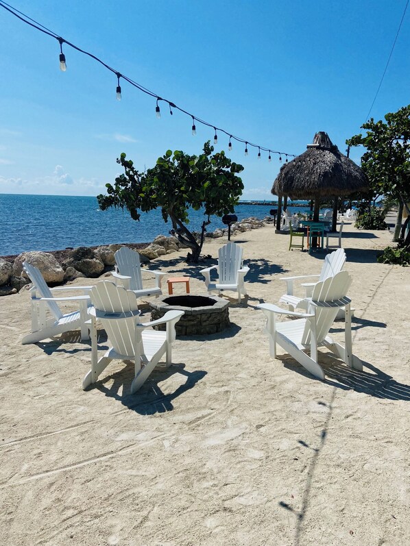 A beachside seating area with white Adirondack chairs arranged around a stone fire pit, overlooking the ocean.