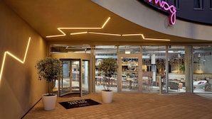 A modern hotel entrance with a neon sign, glass doors, and potted plants. - Moxy Karlsruhe (Karlsruhe)