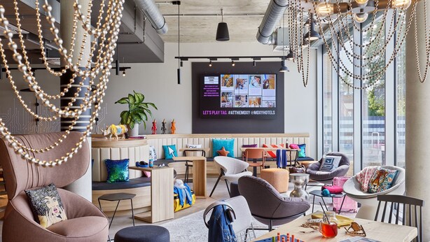 A modern hotel lobby with a large TV screen displaying a promotional video, comfortable seating areas, and a large window offering a view of the city. - Moxy Karlsruhe (Karlsruhe)