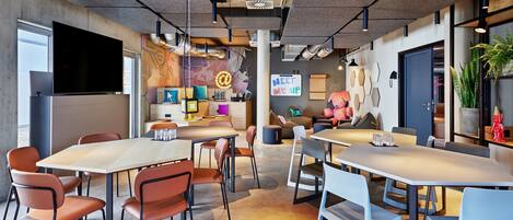 A modern office space with multiple tables and chairs, a large flat-screen TV, and a colorful mural on the wall.