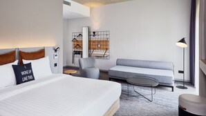 A modern hotel room with a large bed, a grey sofa, a round coffee table, and a chair. There is a wardrobe with a mirror and a hanging rack. - Moxy Karlsruhe (Karlsruhe)