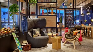 A modern lounge area with a comfortable seating arrangement, a small wooden table, and a view of the cityscape through large windows. - Moxy Karlsruhe (Karlsruhe)