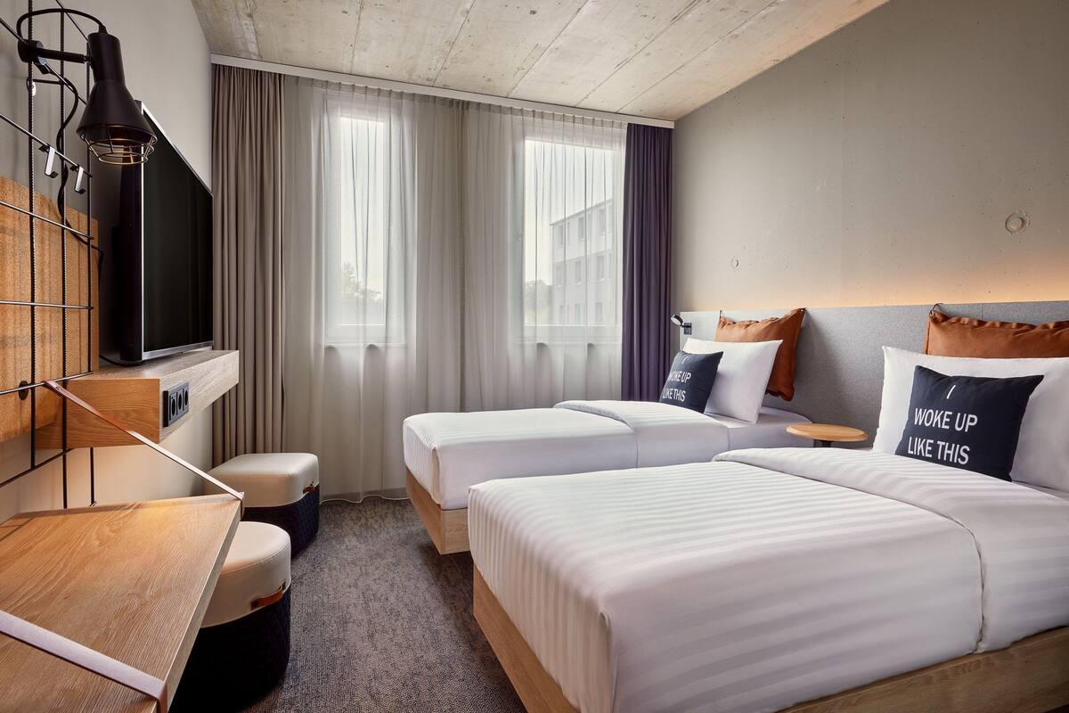 A hotel room with two beds, a desk, a TV, and a window with curtains.