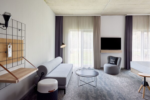 Una habitación de hotel moderna con un sofá gris, una mesa redonda, un sillón gris, un televisor montado en la pared y una ventana con cortinas.