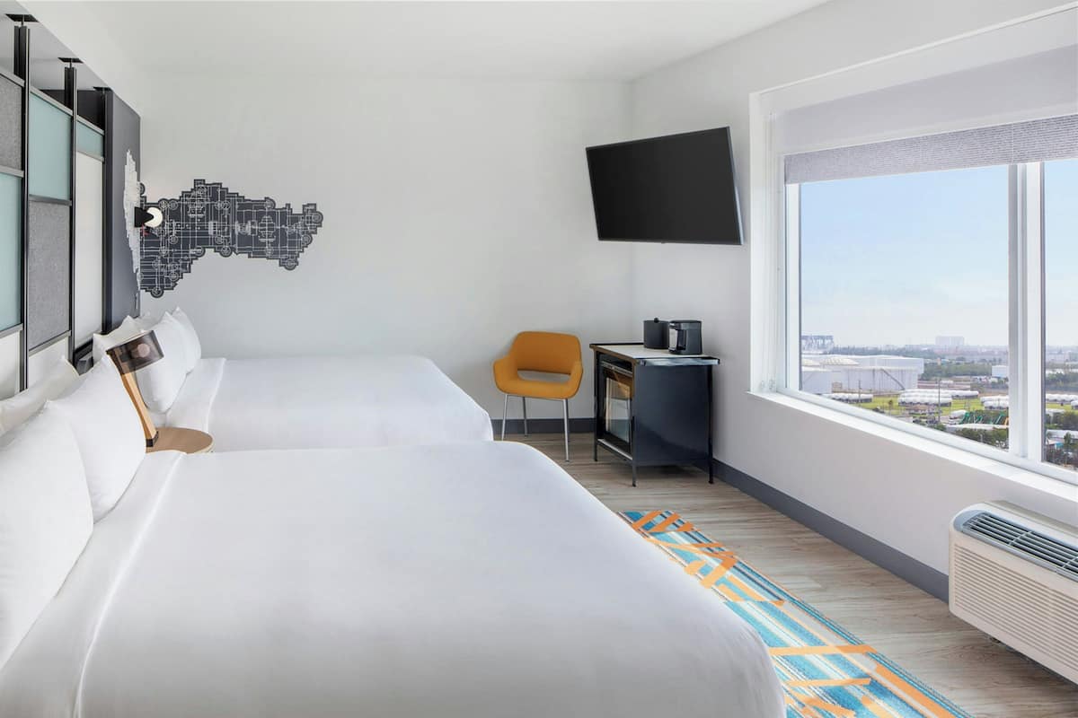Ein Hotelzimmer mit zwei Betten, einem Schreibtisch, einem Stuhl, einem Flachbildfernseher und einem großen Fenster mit Blick auf die Stadt.