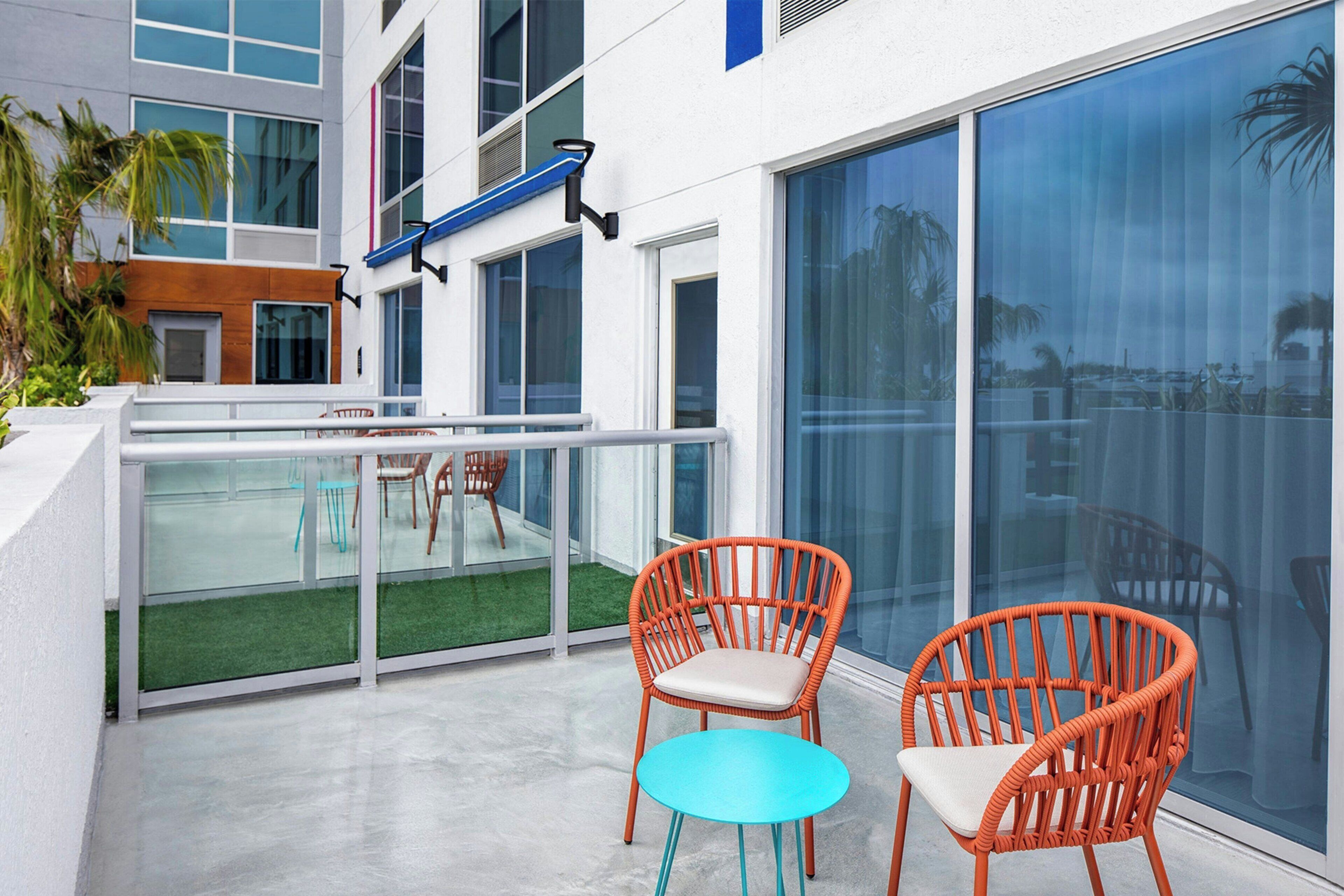 A modern outdoor balcony with two wicker chairs and a small table, overlooking a pool and a building with large windows.