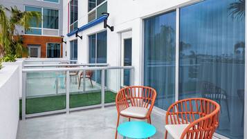 Un balcón moderno al aire libre con dos sillas de mimbre y una pequeña mesa, con vistas a una piscina y un edificio con amplios ventanales.