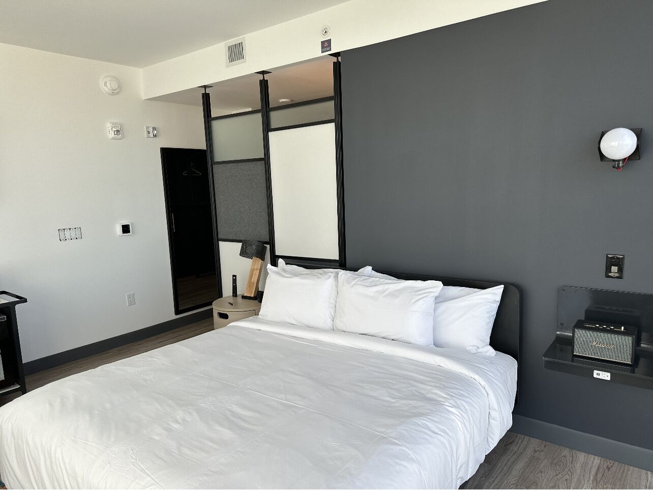 A modern hotel room with a large bed, a bedside table, a lamp, and a wall-mounted speaker.