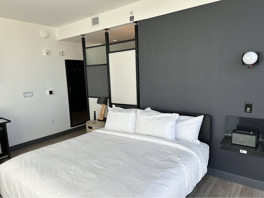 A modern hotel room with a large bed, a bedside table, a lamp, and a wall-mounted speaker.