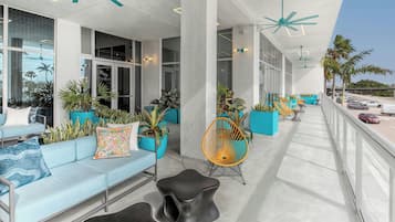 Een moderne buitenzitplek met een lichtblauwe bank, een zwarte kruk en een gele stoel. Er staan potplanten en loopt een leuning langs de rand.