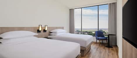 A hotel room with two beds, a large window with a city view, a desk with a chair, and a flat-screen TV.