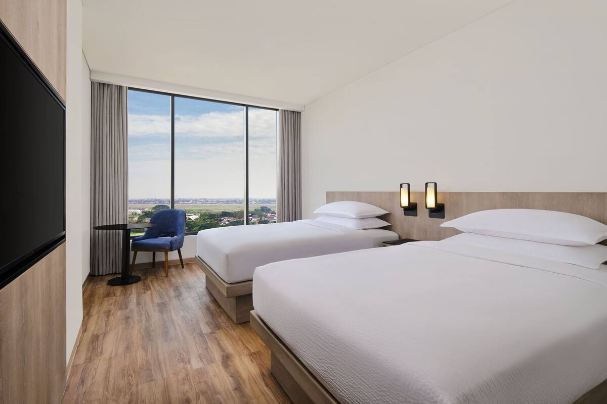 Quarto de hotel com duas camas, uma janela grande com vista, uma escrivaninha com cadeira e uma televisão.