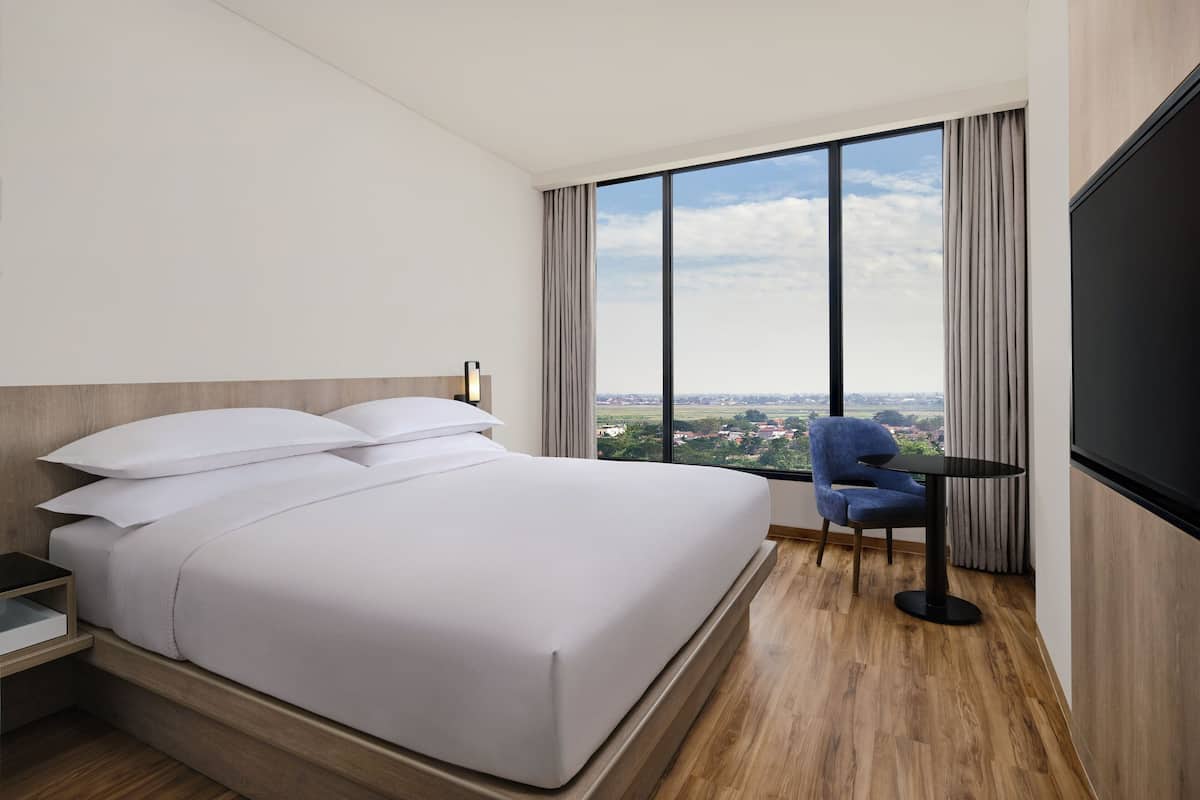 Quarto de hotel com uma cama grande, uma mesa com cadeira, televisão e vista para a cidade.