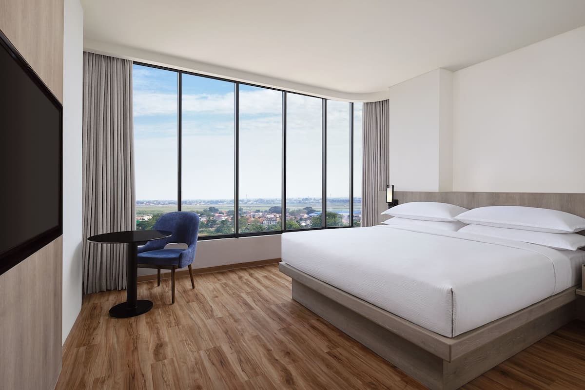 Quarto de hotel moderno com uma cama grande, uma poltrona, televisão e uma janela ampla com vista para a cidade.