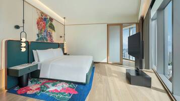 A modern hotel room with a large bed, a flat-screen TV, a floral rug, and a city view.