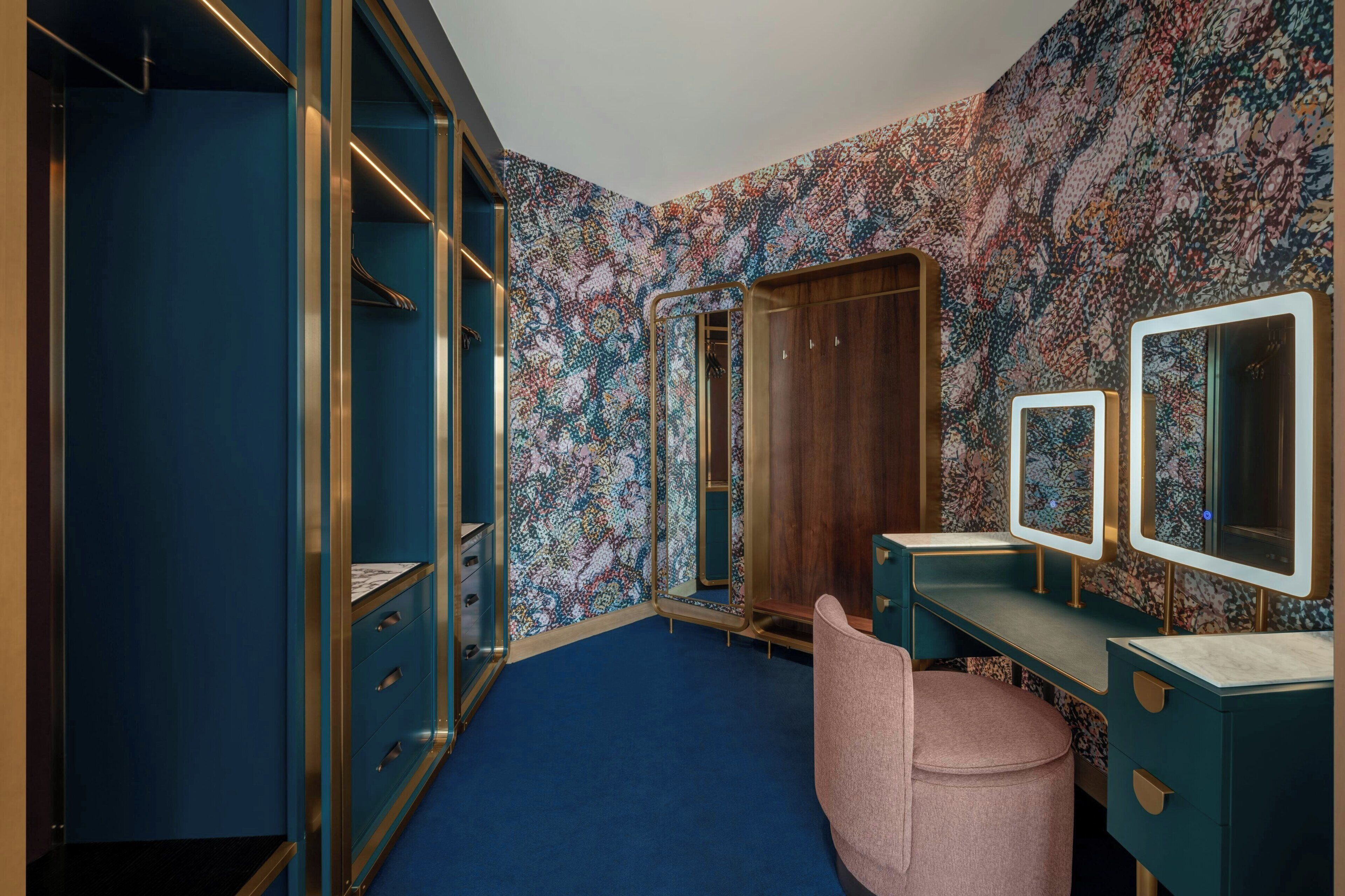 A room with a blue carpet, a pink chair, a wooden wardrobe, and a vanity with mirrors.