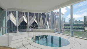 A modern indoor pool with a glass barrier, large windows offering a city view, and a geometric wall design. - W Macau - Studio City (Cotai)