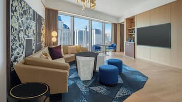 A modern hotel room with a sofa, ottomans, a coffee table, and a large TV. The room has a city view through large windows.