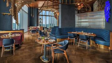 A modern restaurant with a wooden herringbone floor, blue upholstered seating, and large windows offering a view of the city.