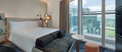 Una habitación de hotel moderna con una cama grande, una zona de estar, una mesita pequeña y un ventanal con vistas a la ciudad.