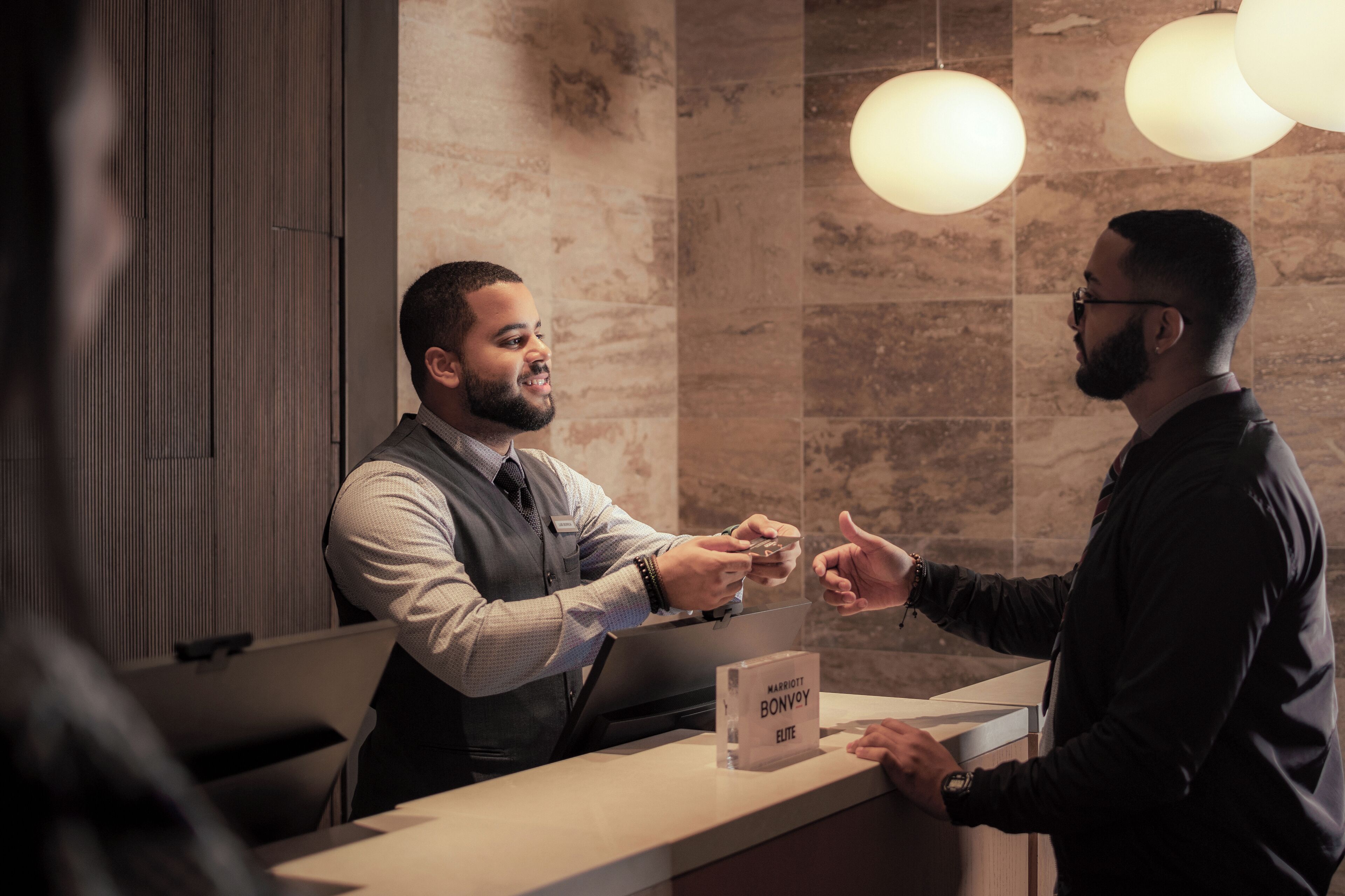 Uma área de recepção de hotel com parede de mármore e iluminação pendente. Uma placa indica que é um empreendimento Marriott Bonvoy Elite.