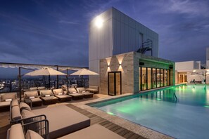 Un área de piscina en la azotea con sillones y sombrillas, un edificio moderno con fachada de vidrio y vistas a la ciudad al atardecer.