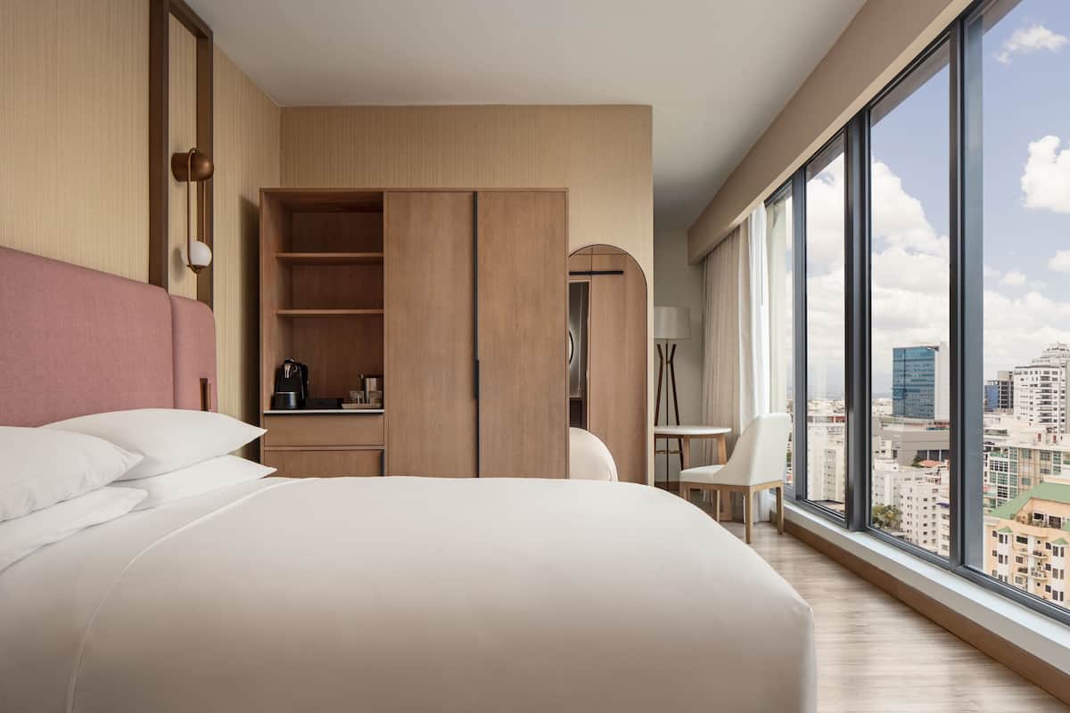 A modern hotel room with a large bed, a wardrobe, a desk, and a view of the city.
