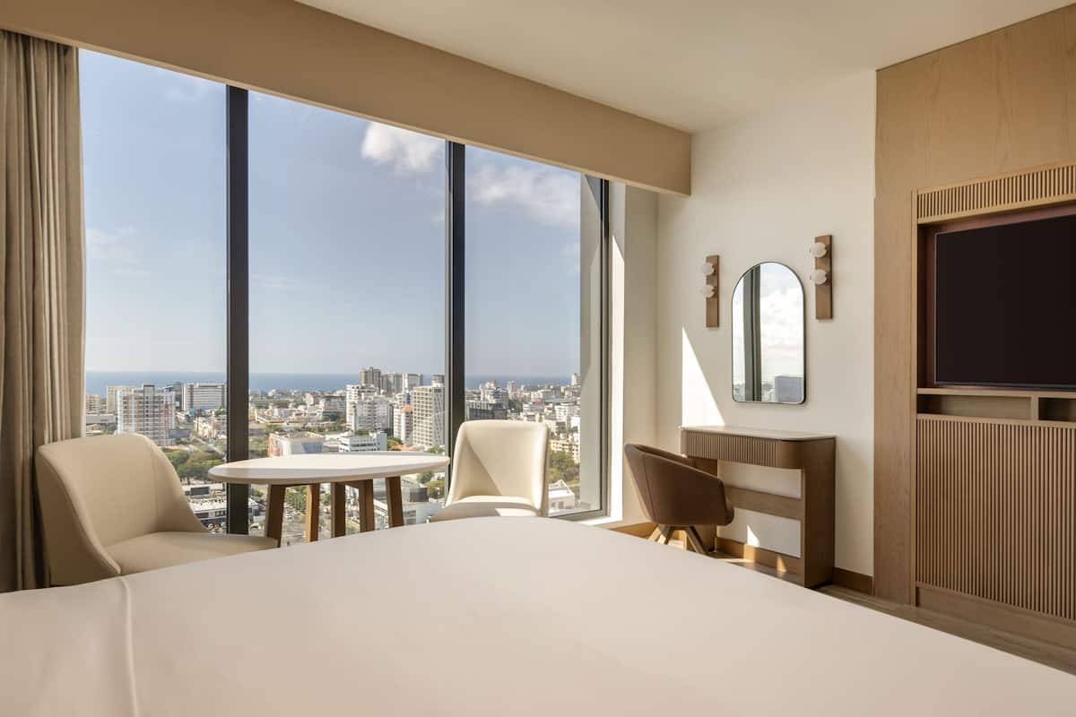 A hotel room with a large bed, a round table, two chairs, a TV, and a city view through large windows.