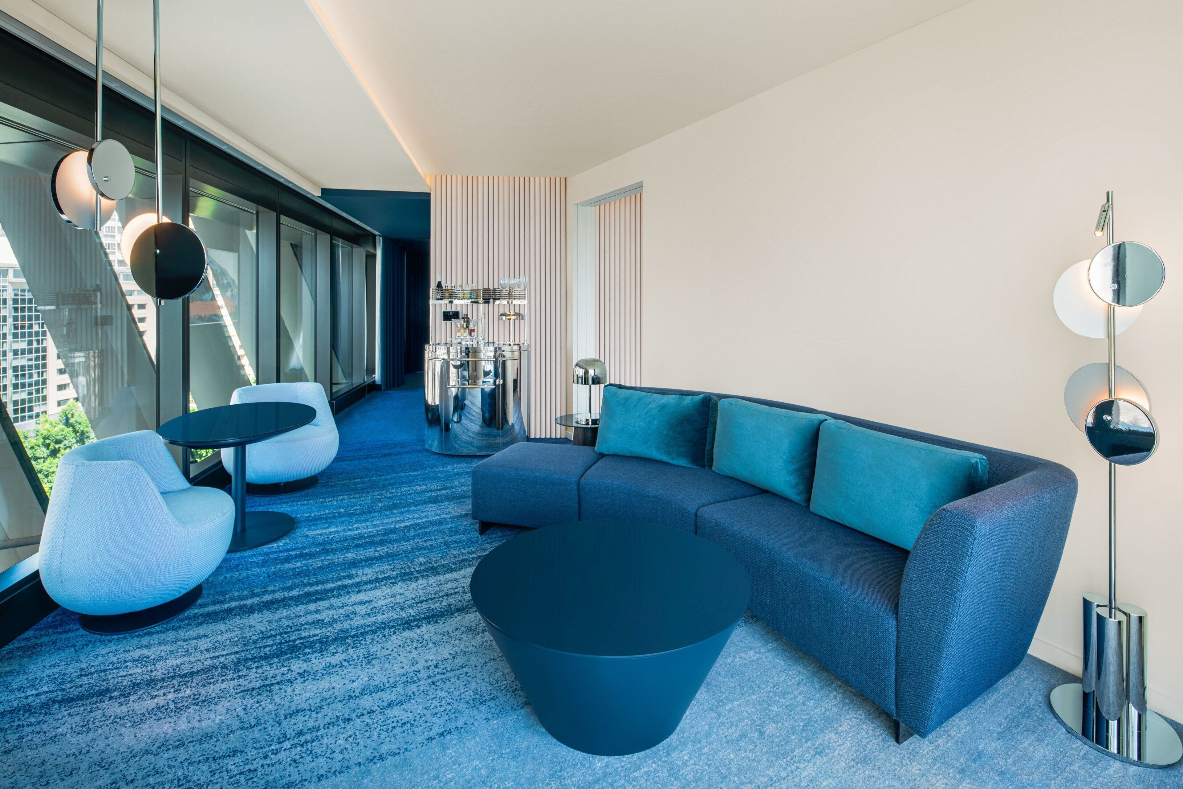A modern living room with a blue sofa, white armchairs, a dark coffee table, and a large window offering a city view.