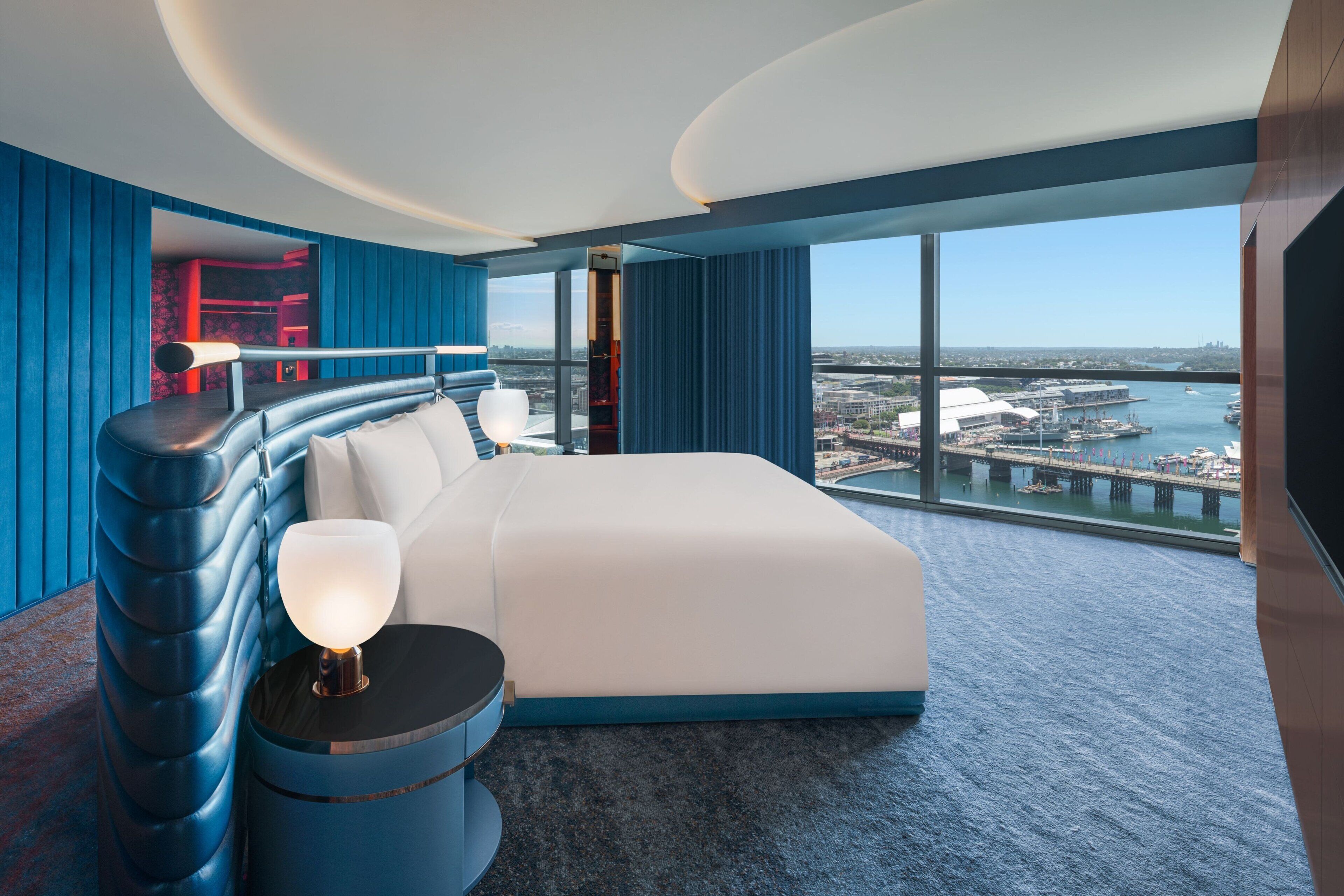A modern hotel room with a large bed, a bedside table with a lamp, a view of the city and water through large windows.
