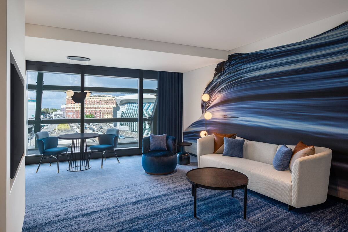 A modern hotel room with a large window, a round table, two chairs, a sofa, and a coffee table.