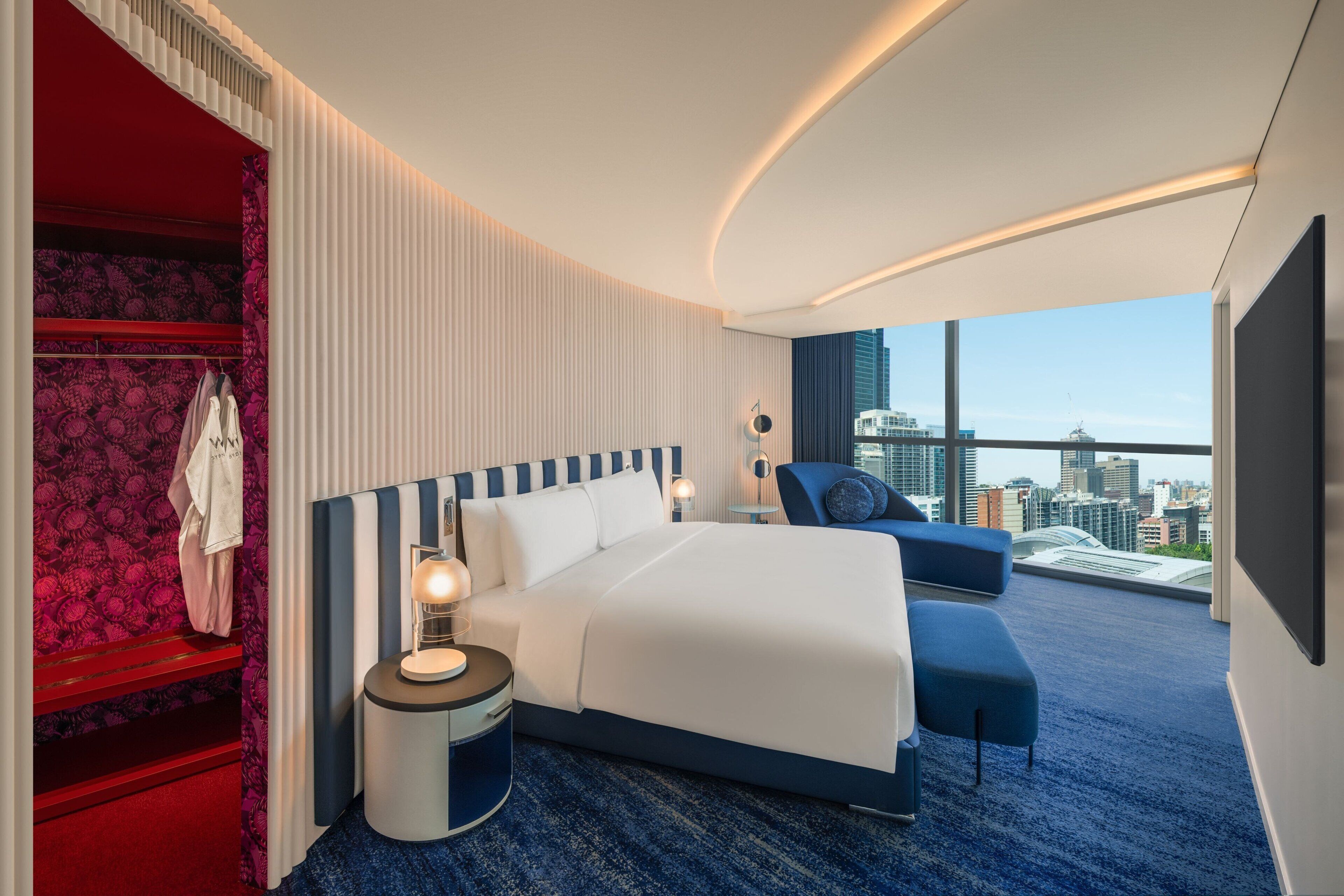 A modern hotel room with a large bed, a seating area, a wardrobe, and a city view.