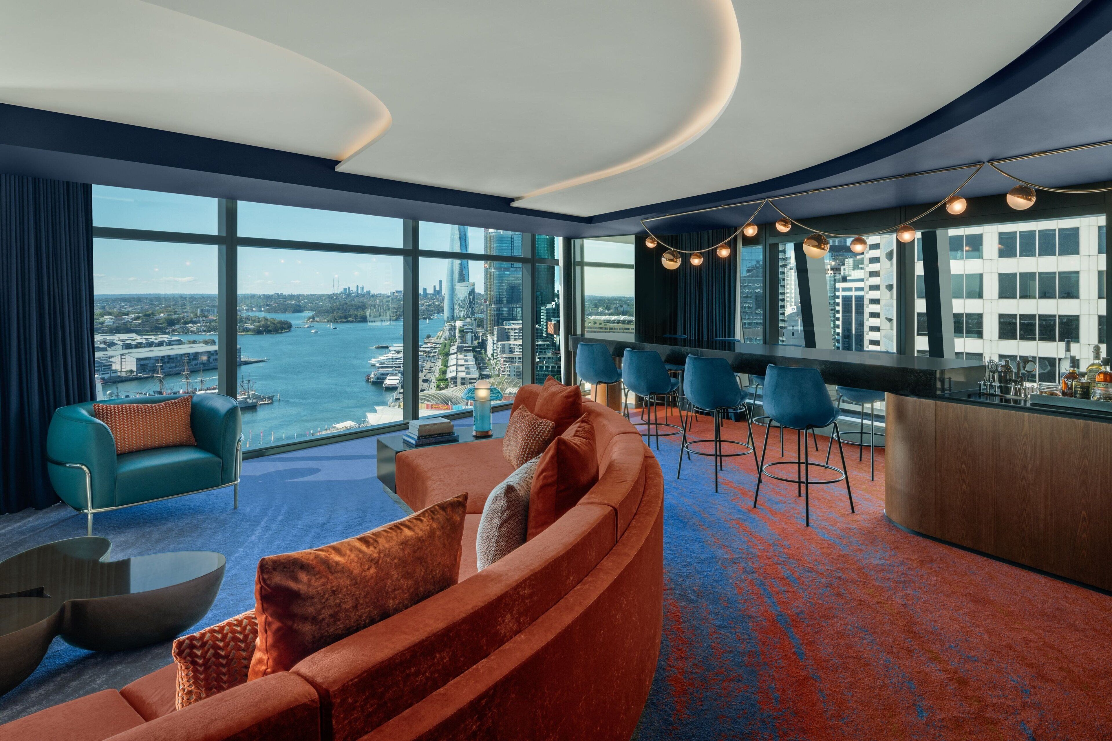 Una habitación de hotel moderna con un sofá curvo, una zona de bar y ventanas amplias con vistas a la ciudad.