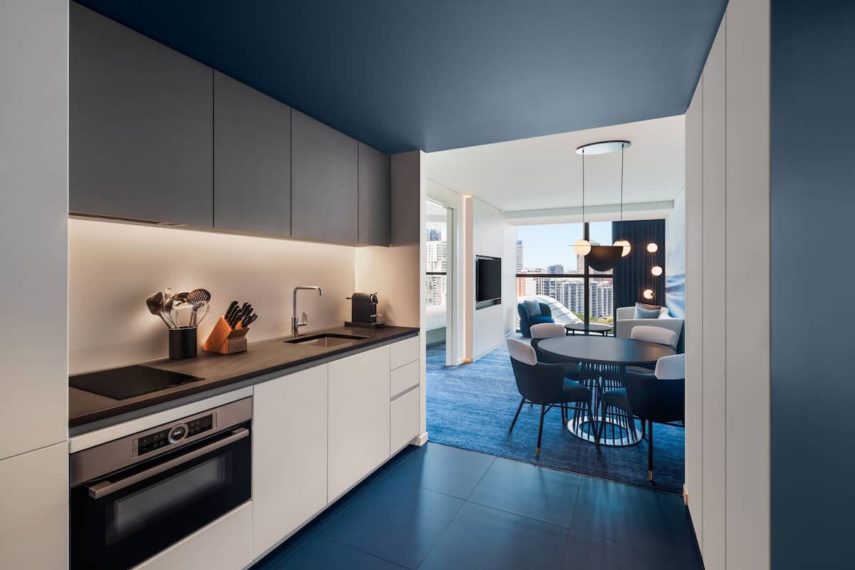 A modern kitchen with integrated appliances, a dining area with a round table and chairs, and a view of the cityscape through large windows.