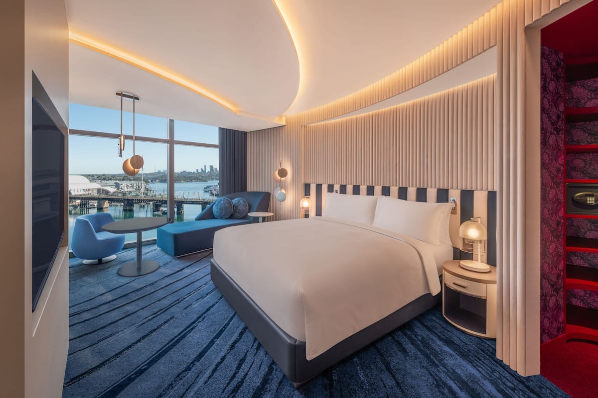 A modern hotel room with a large bed, a seating area with blue chairs, a TV, and a view of a cityscape through large windows.