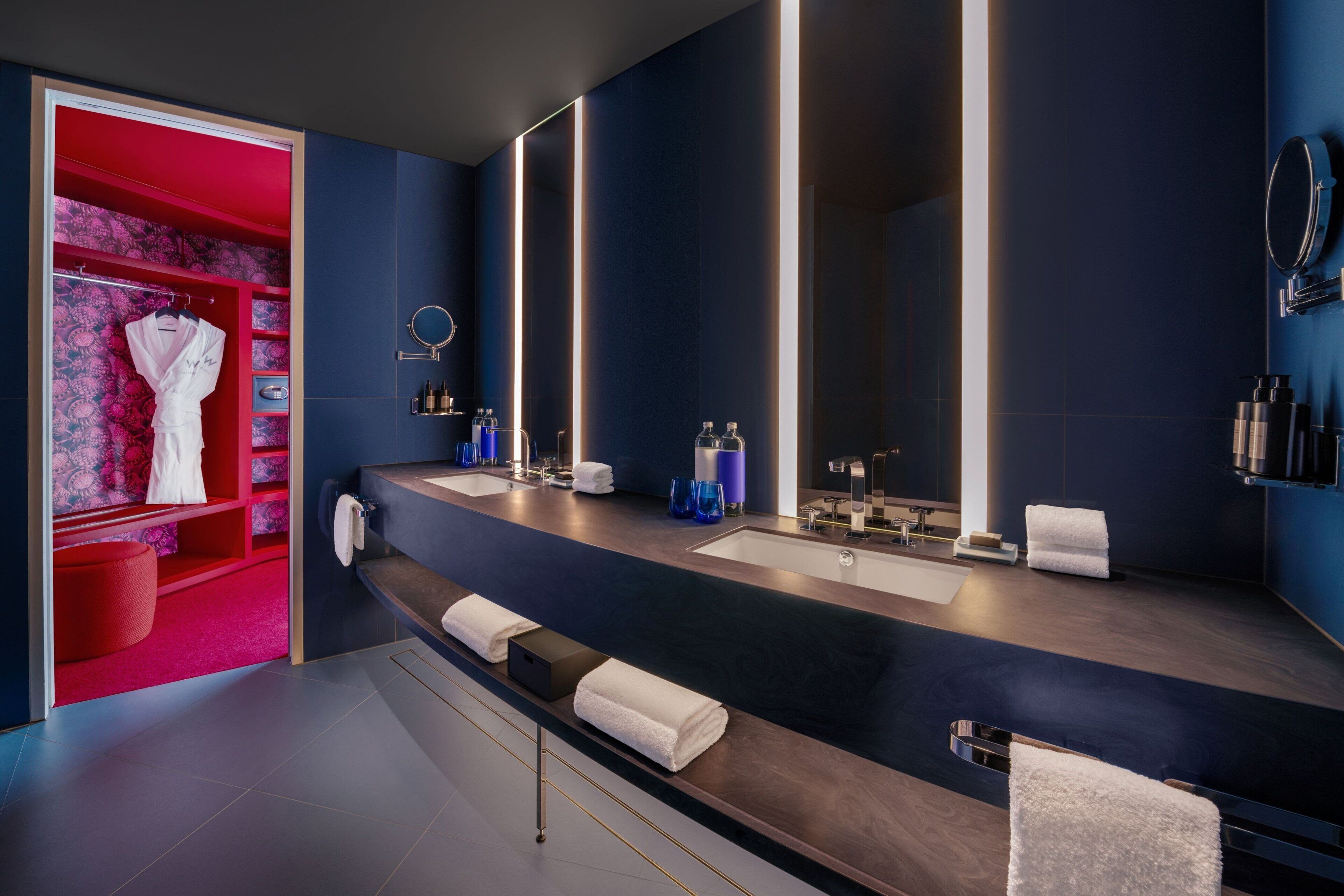 A modern bathroom with a dark blue wall, a large mirror, two sinks, and a red vanity area with a robe and towel rack.