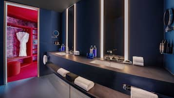 Un baño moderno con pared azul oscuro, un espejo grande, dos lavamanos y una zona de tocador roja con perchero para bata y toallas.