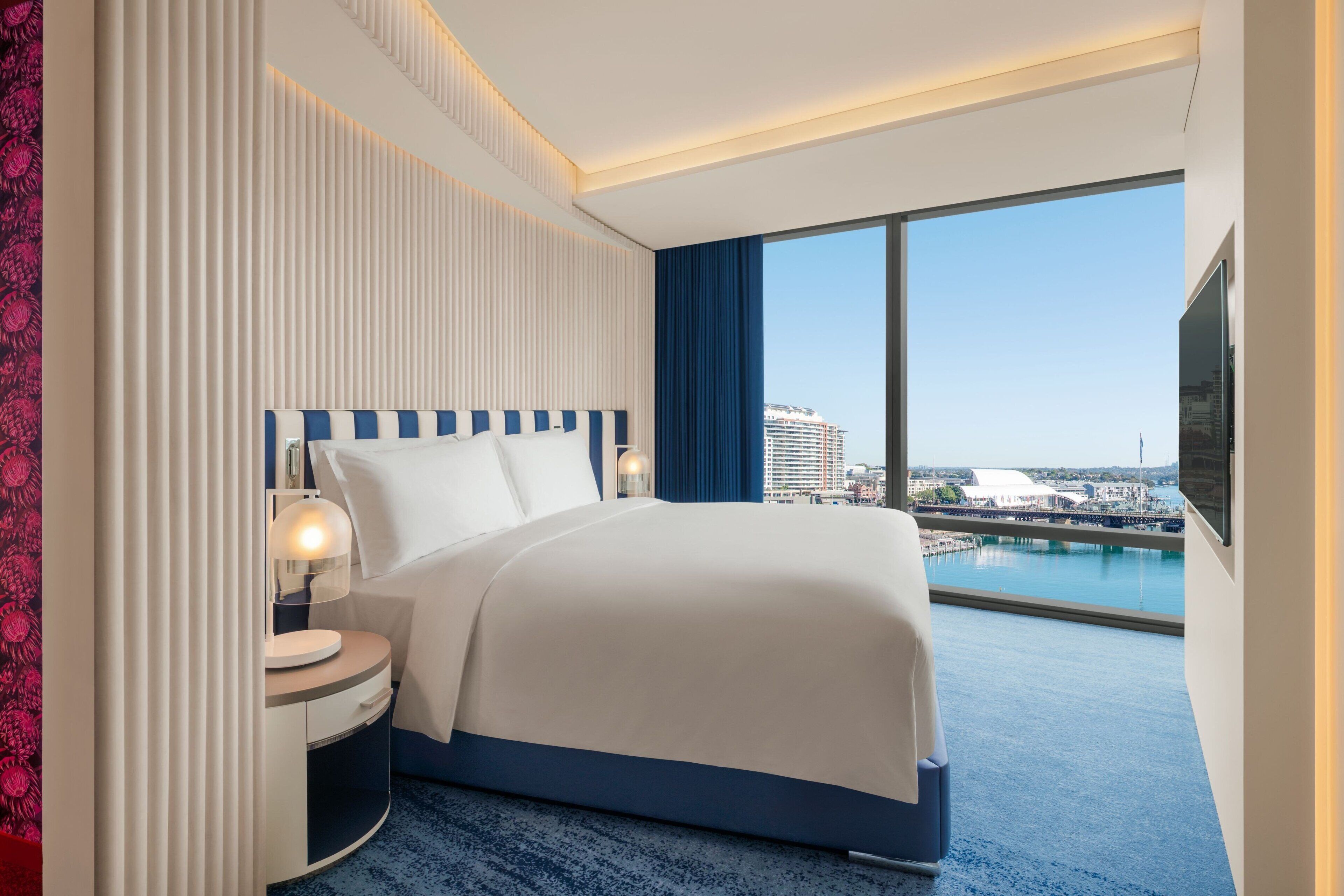 A modern hotel room with a large bed, a nightstand, a lamp, and a view of a marina and buildings.
