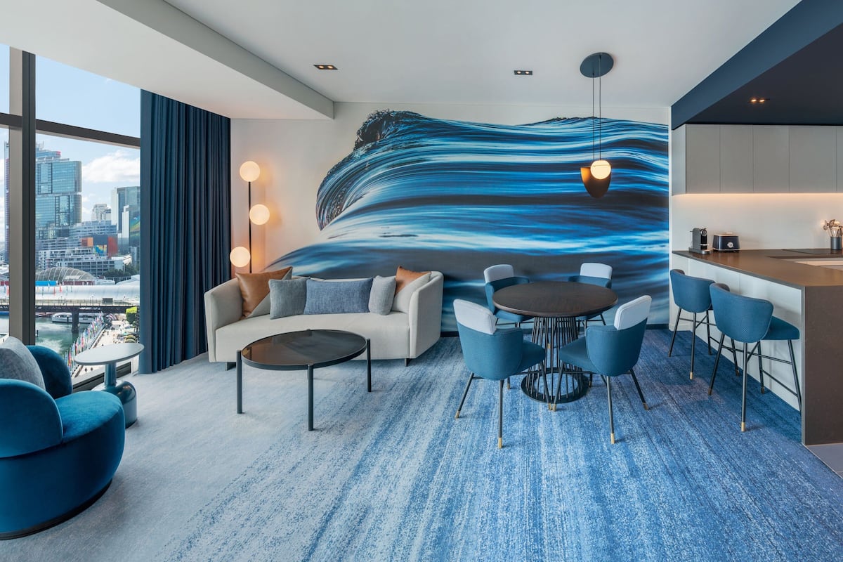 A modern hotel room with a large city view, a wave-themed mural, a cozy seating area, and a kitchenette.