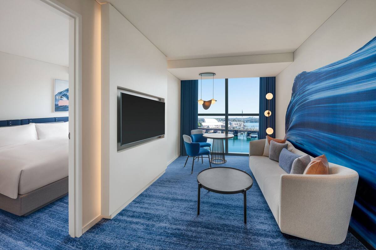 A modern hotel room with a bed, a seating area with a sofa and chairs, a flat-screen TV mounted on the wall, and a large window offering a view of the city.