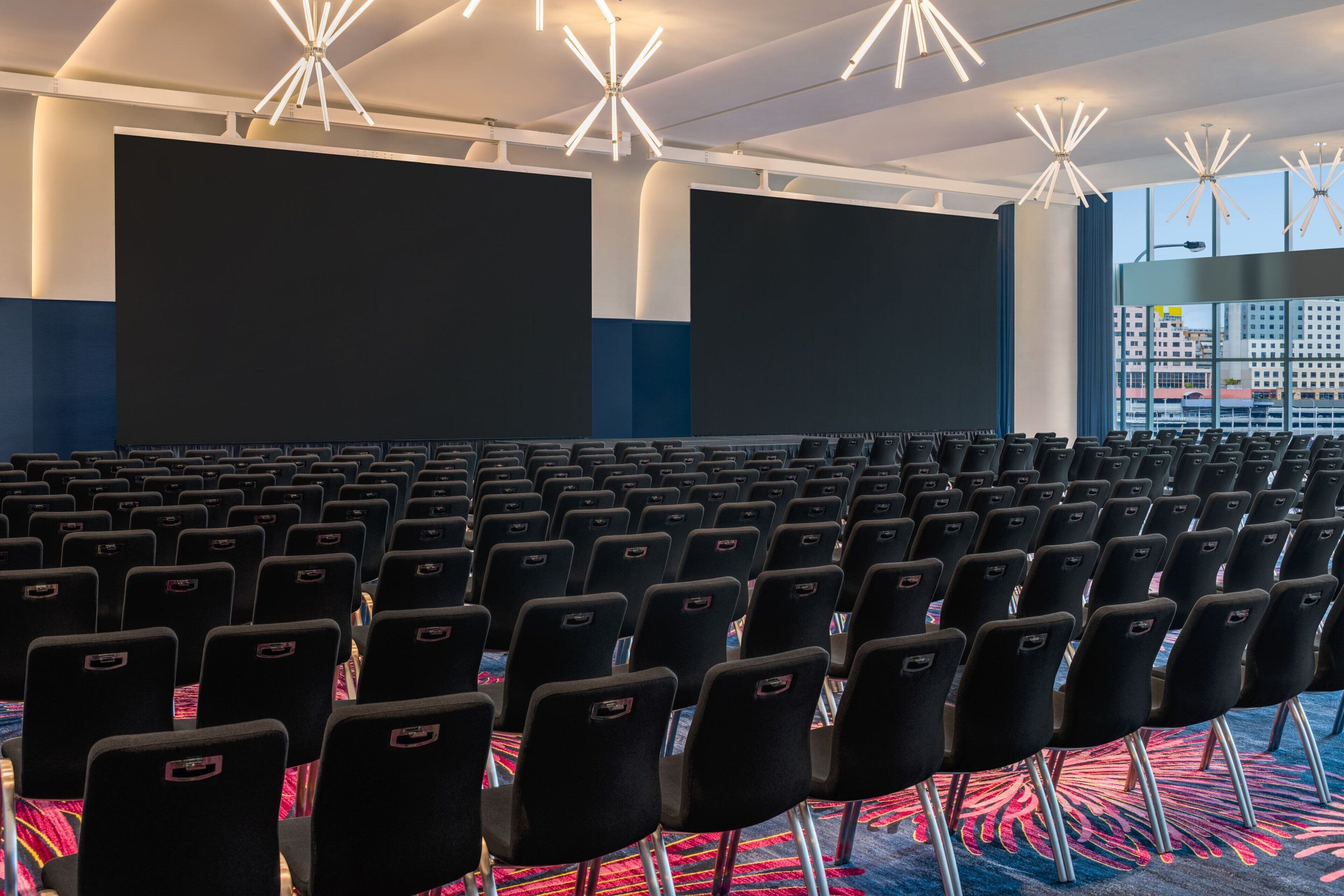A large indoor auditorium with rows of black chairs and large screens.