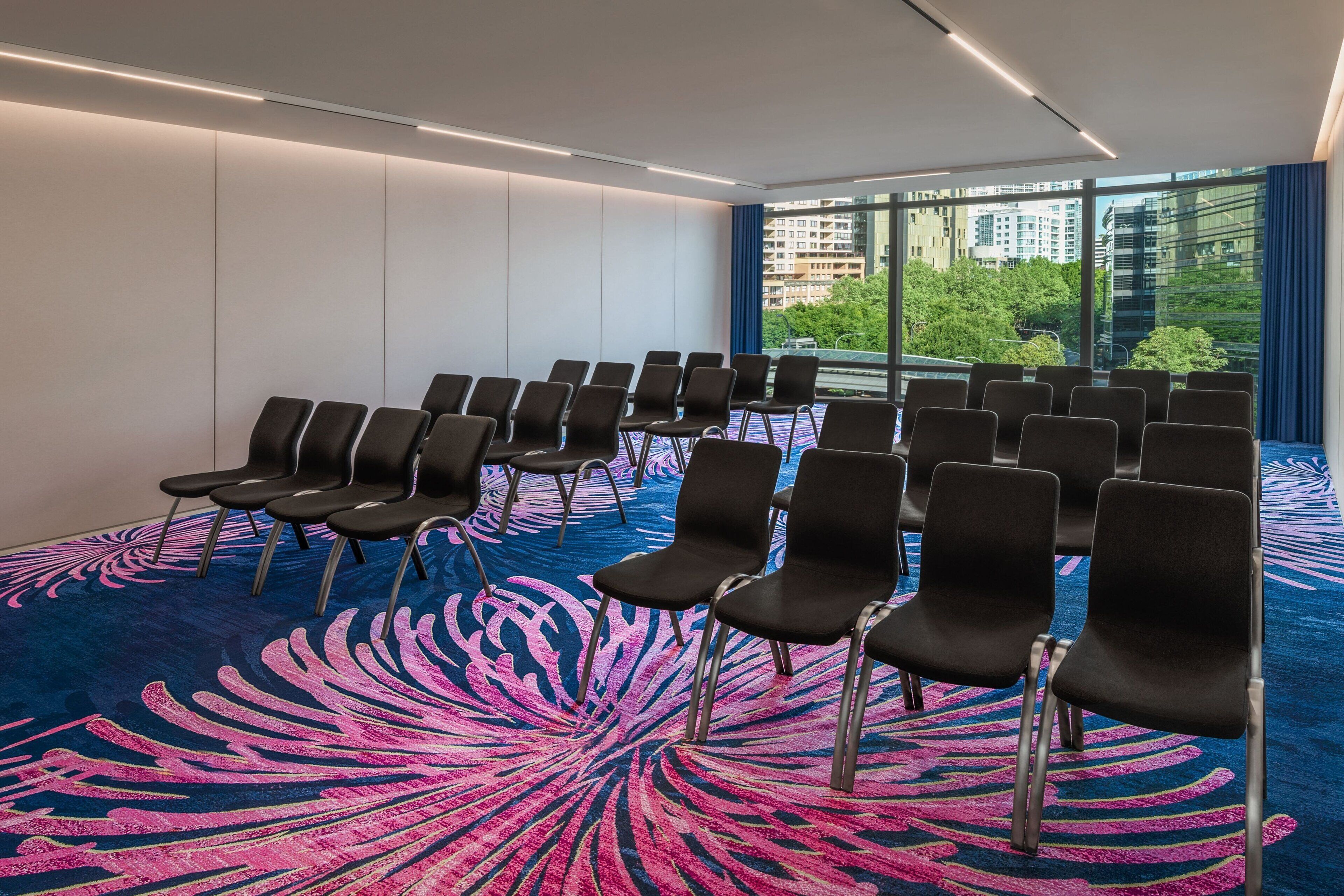 Un salón de conferencias con filas de sillas negras, una alfombra estampada y amplios ventanales con vistas a edificios y árboles.
