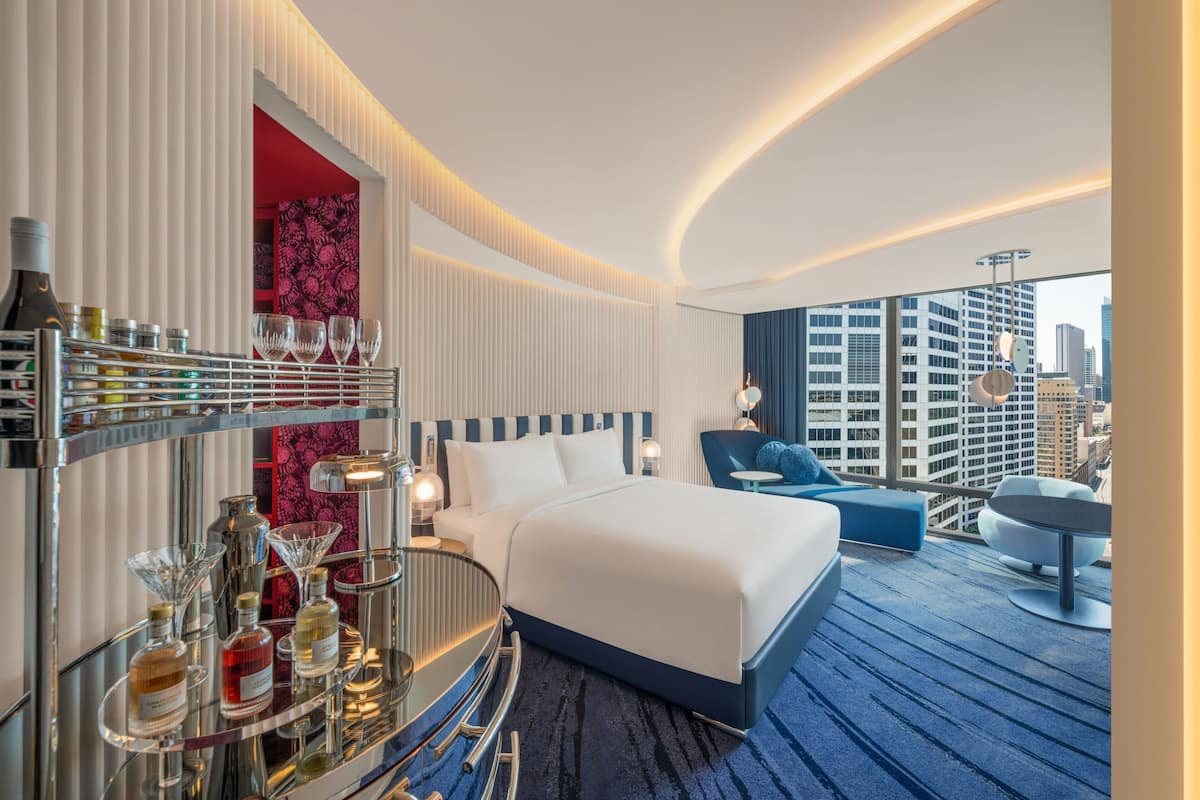 A modern hotel room with a large bed, a seating area with a view of the city, and a bar area with bottles and glasses.