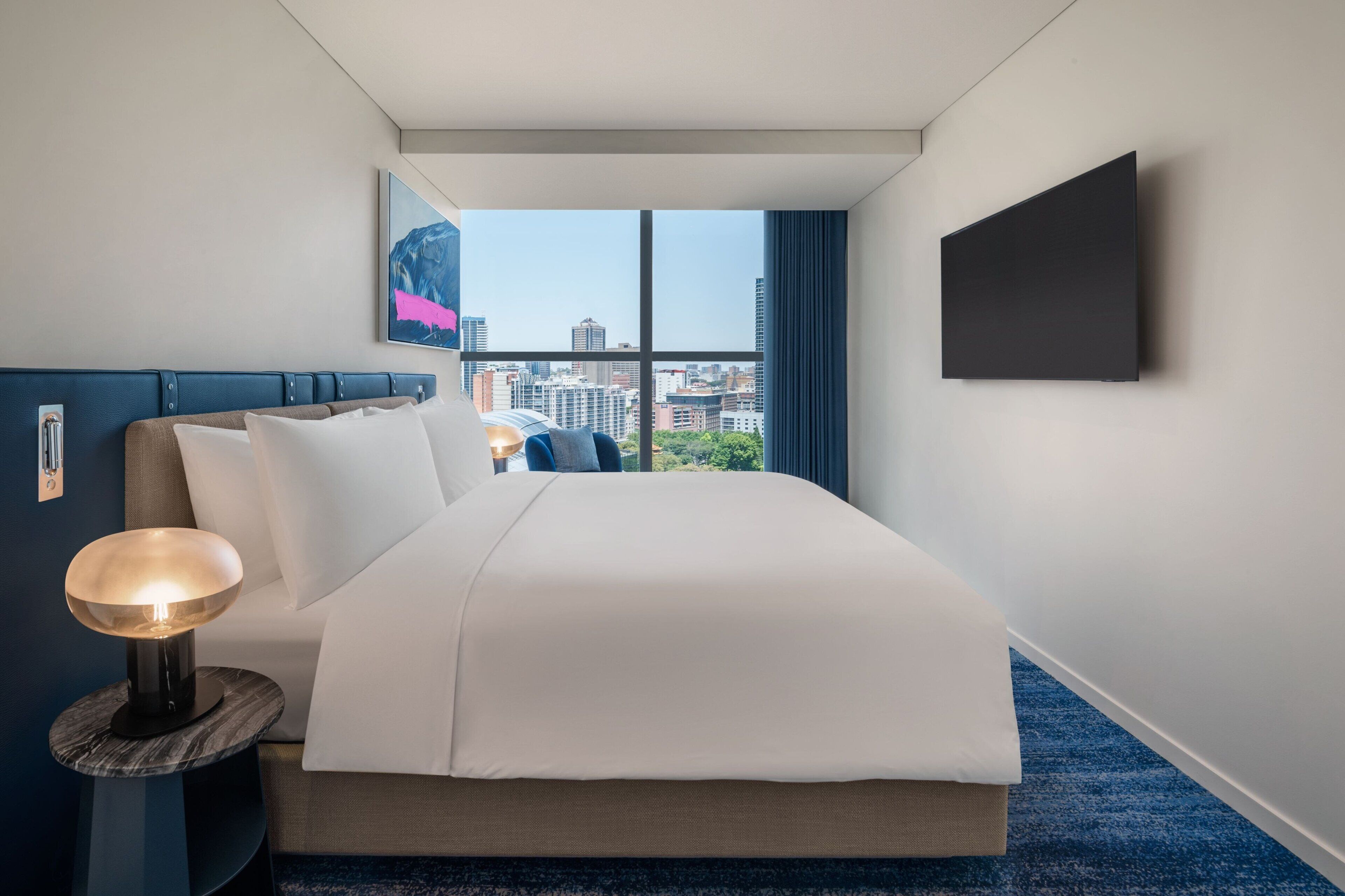 A hotel room with a large bed, a bedside lamp, a flat-screen TV, and a city view through the window.