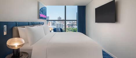 A hotel room with a large bed, a bedside lamp, a flat-screen TV, and a city view through the window.